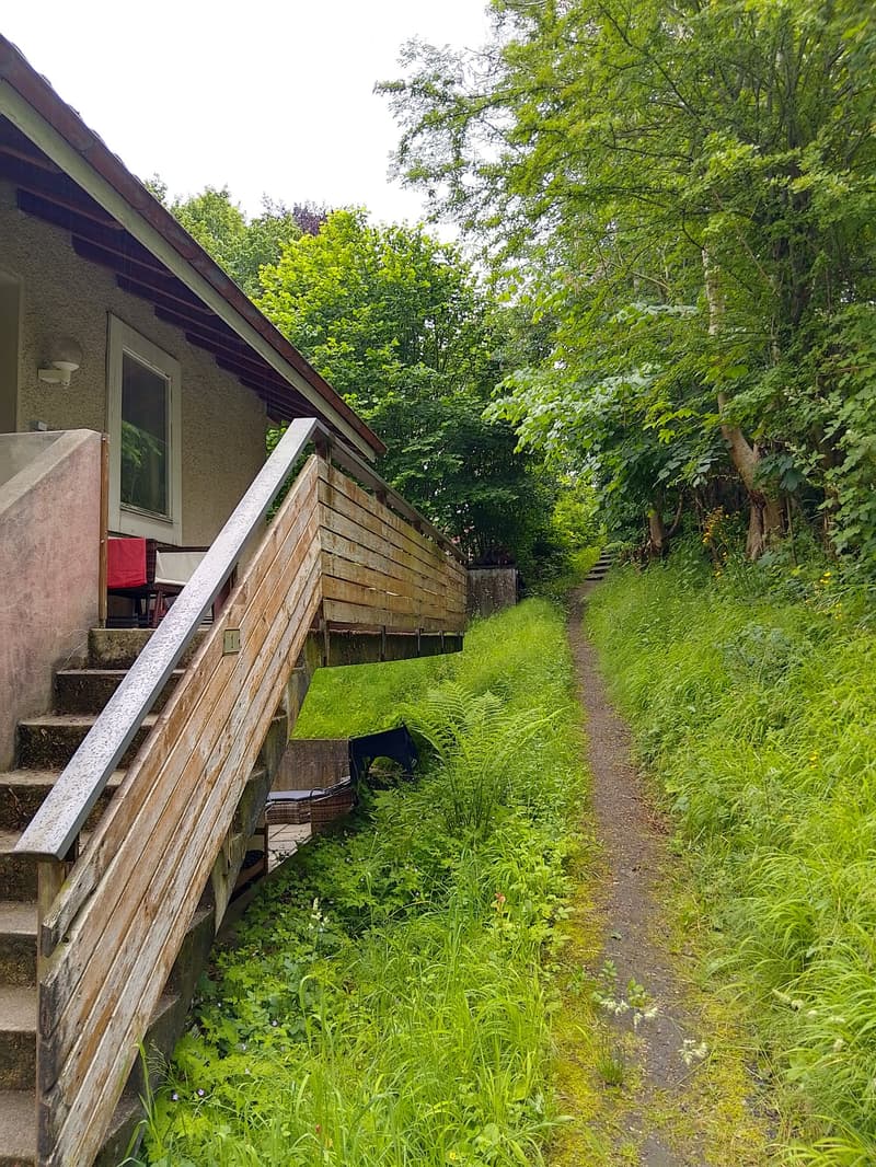 A wooden staircase leads up to a house with a small porch area, surrounded by lush greenery and a narrow pathway.