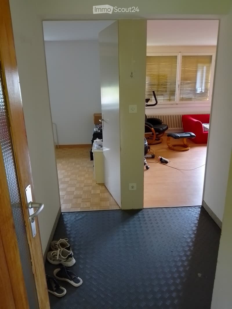 A narrow hallway with a dark blue patterned floor, leading to a room with wooden flooring, a red sofa, a black chair, and exercise equipment. A pair of shoes is placed on the hallway floor near the door.