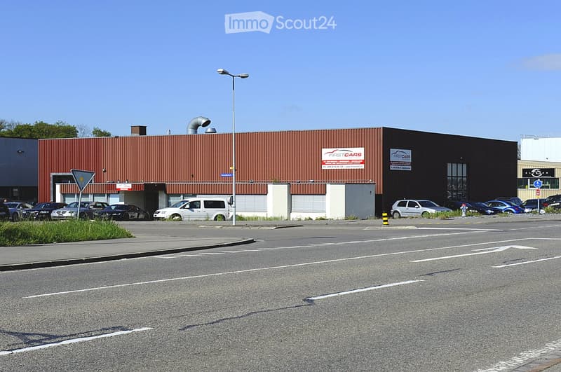 A large industrial-style building with a red metal roof and signage for "FIRSTCARS" visible. The building has multiple entrances and a parking area with several cars.