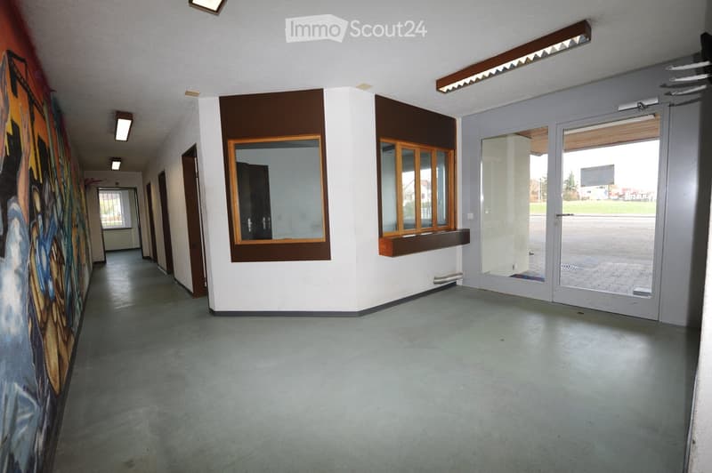 Empty hallway with white walls, grey concrete floor, and large windows with wooden frames. Features a colorful mural on one wall and a glass door leading outside.
