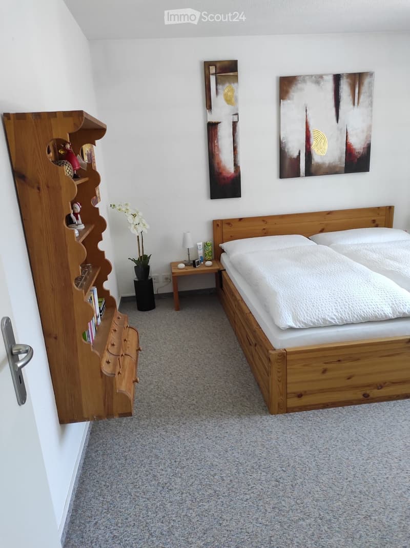 Cozy bedroom with a wooden bed, nightstand, table lamp, wooden shelf with decorative items, and artwork on the walls.