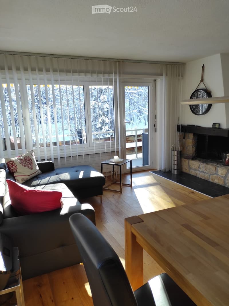 Bright living room with wooden flooring, large windows, a stone fireplace, and a view of snowy trees outside. Features a black leather sofa with colorful cushions, a small side table, and a dining table with chairs.