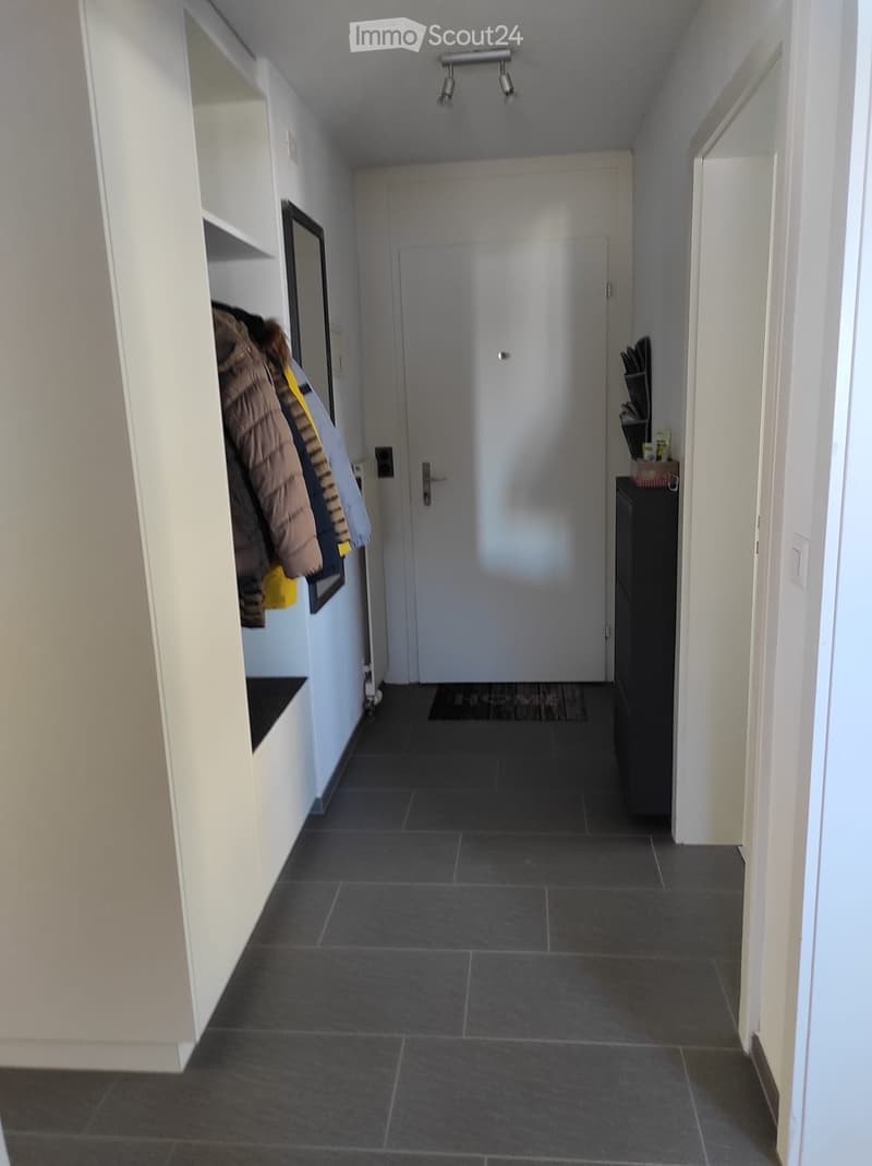 Clean hallway with dark tiled floor, white walls, built-in shelf on left, coat rack with jackets, small table with items on right, closed door at end.