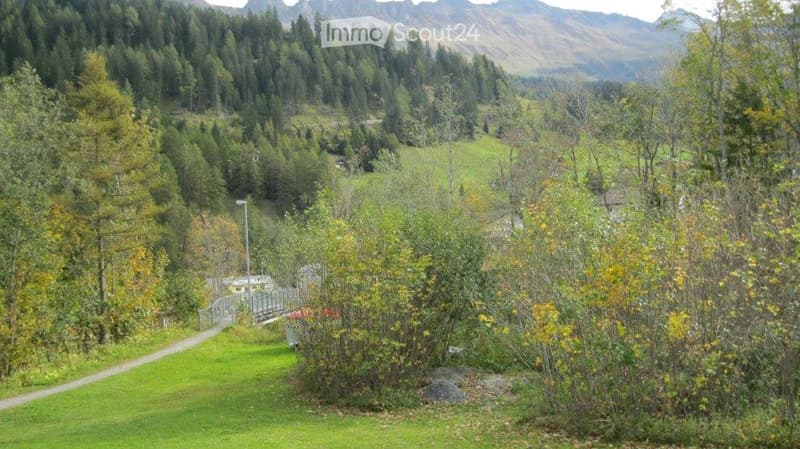 A lush, green garden area with dense trees, shrubs, and a small bridge leading to a pathway. The background features a scenic mountain range.