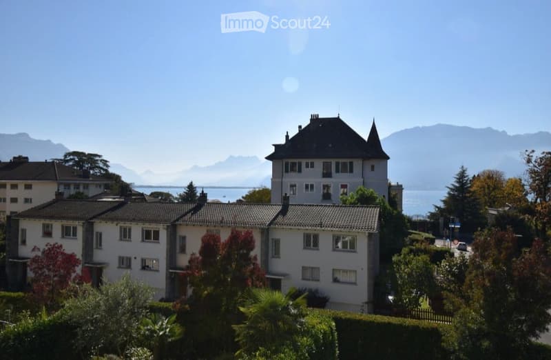A multi-story residential building with a classic architectural style, surrounded by greenery and offering a scenic view of mountains and a body of water in the background.