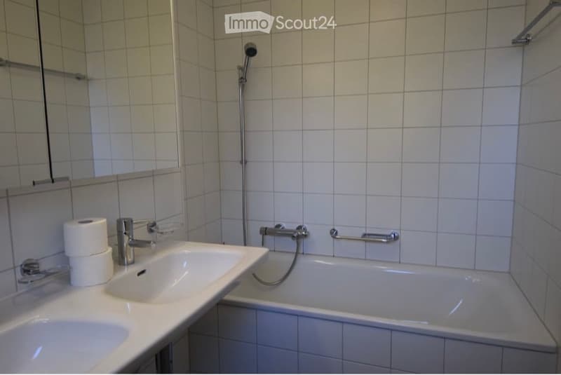 Clean bathroom with white tiles, a sink, a bathtub, a shower head, and a mirror. Toilet paper is placed on the sink counter.