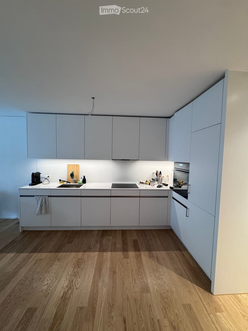 Modern kitchen with light grey cabinets, white countertops, and wooden flooring. Features a sink, faucet, oven, coffee maker, cutting board, and a towel hanging from a cabinet.