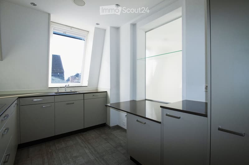 Modern kitchen with grey cabinets, a black countertop, and a sink under a window. The space is bright with natural light from multiple windows, including one with a view of a building exterior.