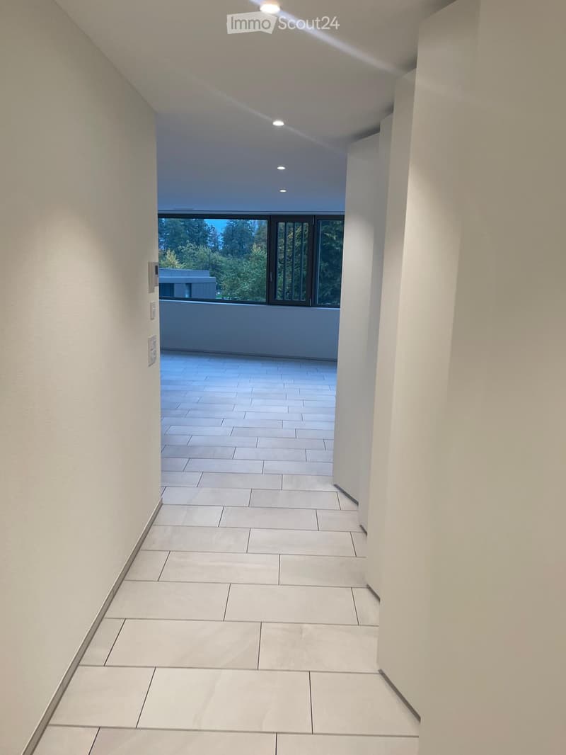 Modern hallway with white tiled flooring, white walls, and recessed lighting. The hallway leads to a balcony area with large windows offering a view of greenery outside.