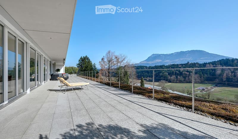Modern balcony with a panoramic mountain view, featuring a lounge chair, glass railing, and large windows.