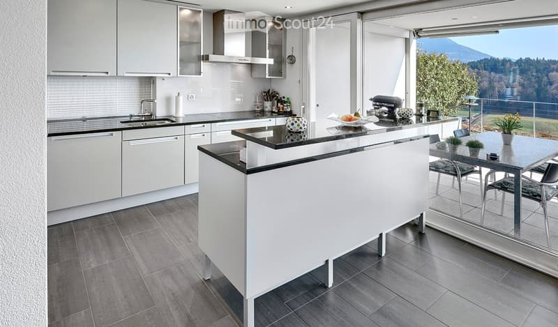 Modern kitchen with white cabinets, black countertops, and a central island. Features a sink, faucet, and various appliances. A dining area with a table and chairs is visible through large windows offering a scenic view.