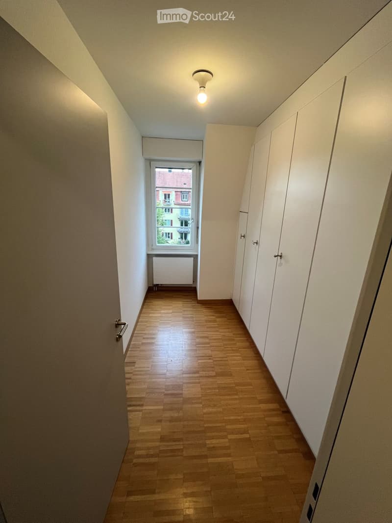 Empty hallway with wooden flooring, white walls, and a window showing a view of a building outside. Features a radiator and built-in storage units.