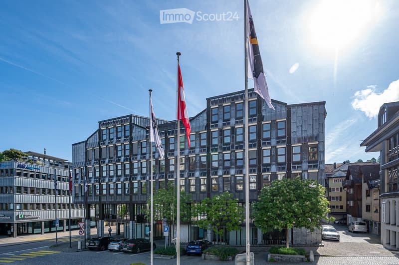 Modern multi-story building with a sleek, dark facade and numerous windows, surrounded by trees and parked cars.