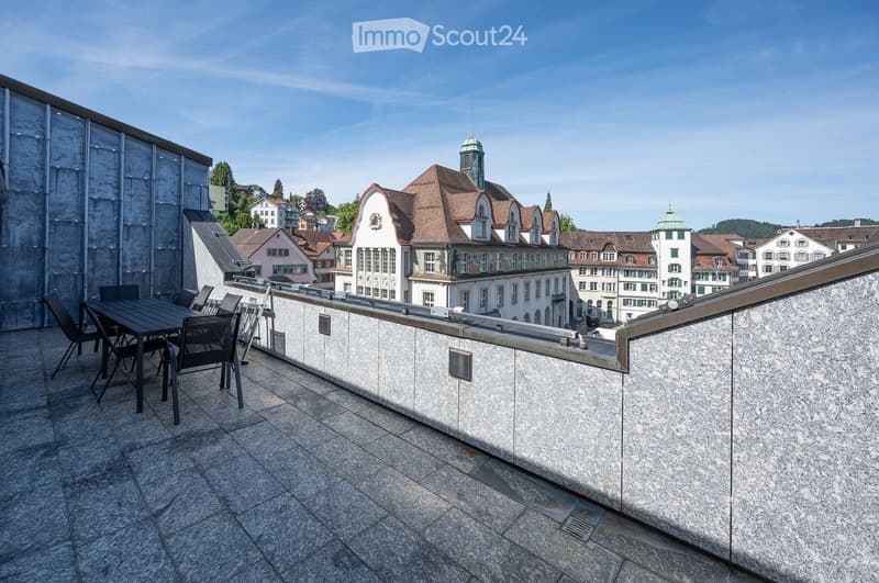 Balcony with a stone floor, a table and chairs, overlooking a cityscape with historic buildings.
