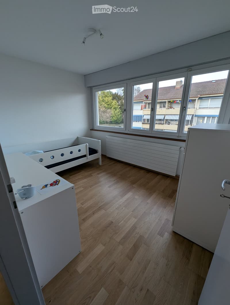 A small, furnished room with wooden flooring, a single bed, a white radiator, and a white cabinet with a cup and a decorative sticker on top.