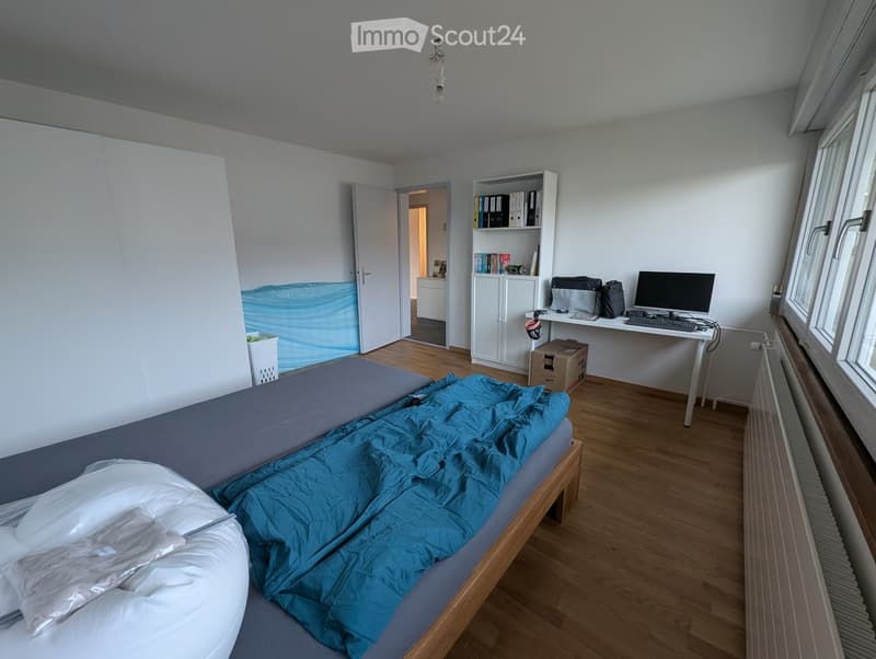 A furnished bedroom with a bed, desk, chair, computer, bookshelf, radiator, and backpack. The room has wooden flooring and white walls.