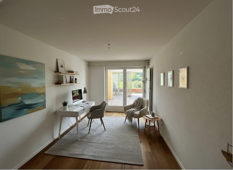 A room with a desk, chair, and a rug. There is a painting on the wall and a side table with a cup. There are also picture frames on the wall.