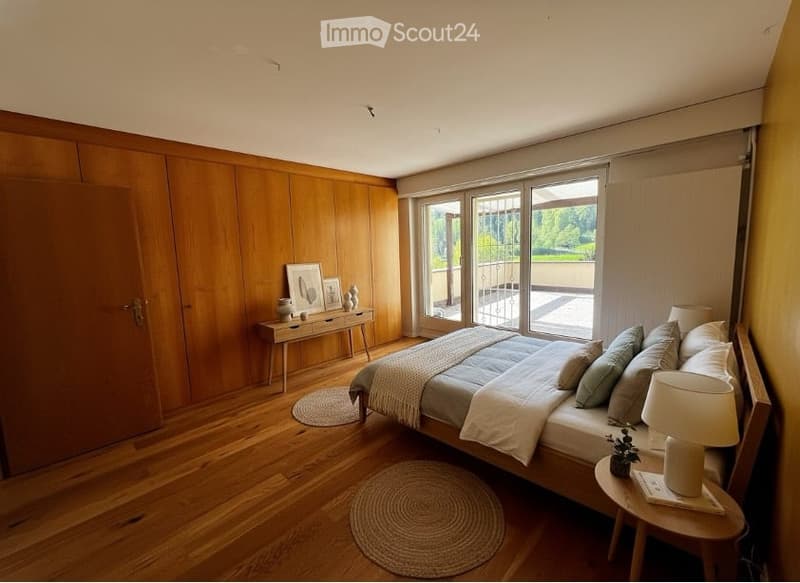 A bedroom with wooden walls and floor, a bed with pillows, a nightstand with a lamp, a rug, and a table with picture frames and vases.