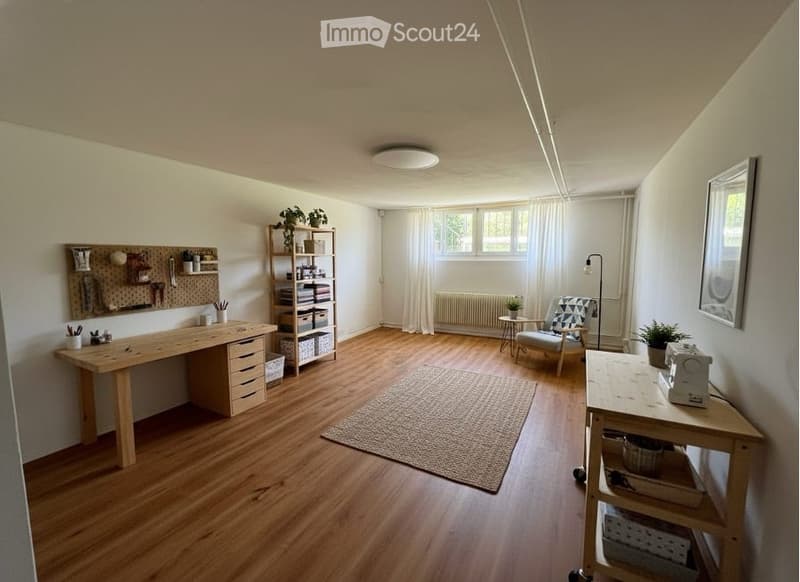A room with wooden floors, white walls, and a ceiling light. It features a desk with drawers, a shelf with a plant, a chair, a lamp, a rug, a table with a sewing machine, and a picture frame on the wall.
