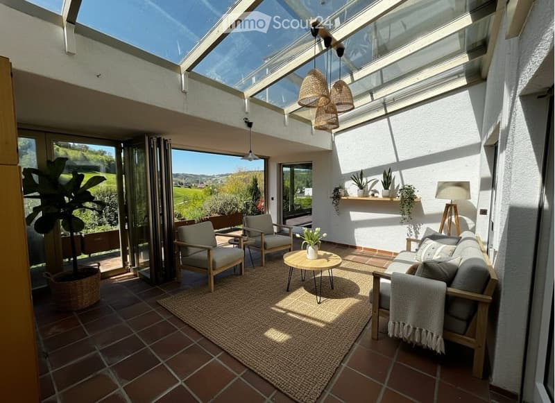 A furnished room with a couch, chairs, coffee table, and a rug. There are plants and lamps hanging from the ceiling. The room has a glass roof and a view of the outside.