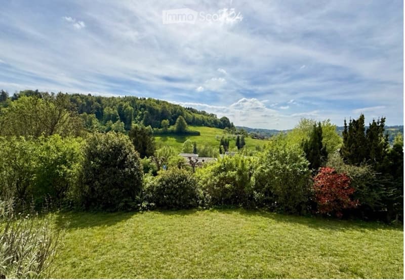 A wide, lush green lawn surrounded by a variety of trees and bushes, with a distant view of a house and a rolling green hill under a partly cloudy sky.