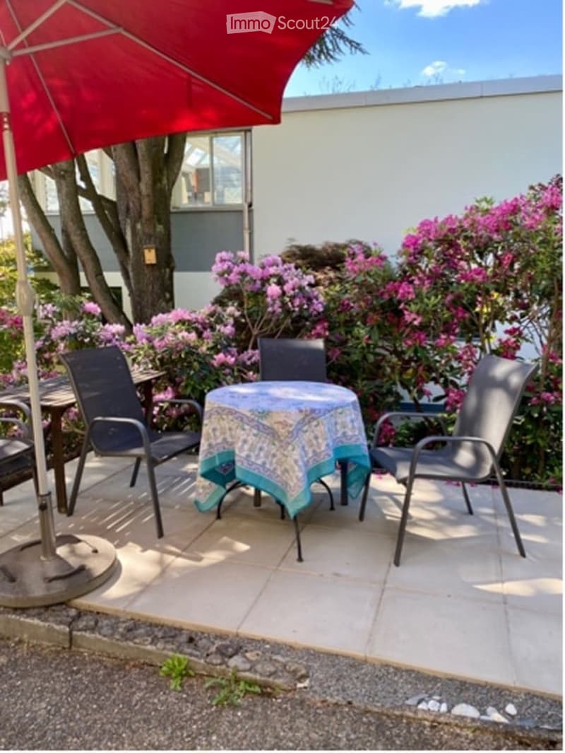 Outdoor seating area with a table and chairs, a red umbrella, and a tablecloth. Surrounded by pink flowers and a tree.