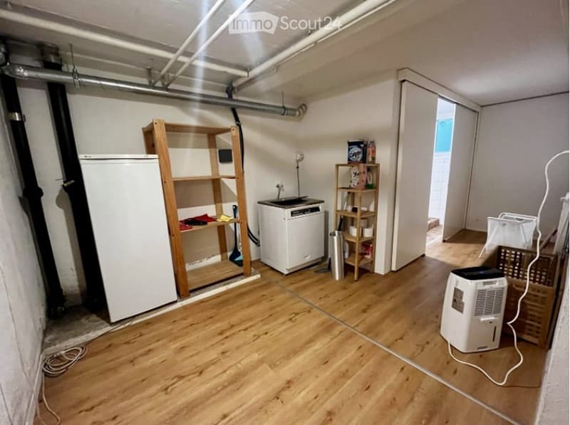 An empty room with wooden flooring and white walls. There are two wooden shelves, a white refrigerator, a white washing machine, a wooden storage box, and a white air conditioner. There are also some pipes and wires on the ceiling.
