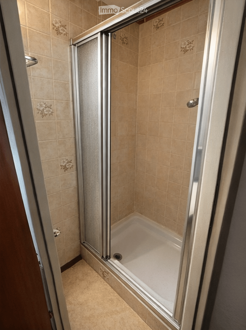 A small bathroom with beige tiled walls and a glass-enclosed shower stall. The shower has a modern, sleek design with a glass door and a built-in seat. The floor is also tiled in a matching beige color.
