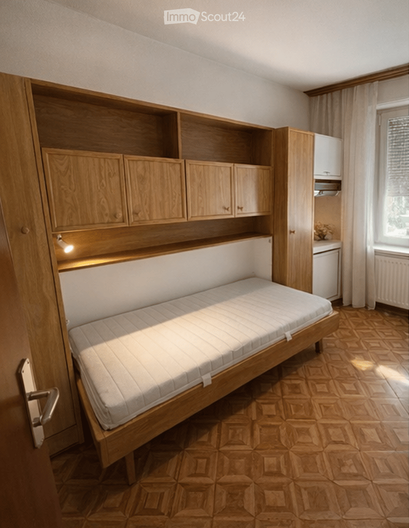 A compact bedroom with a single bed, built-in wooden wardrobe, and small cabinet. The room features wooden flooring and large windows with curtains.