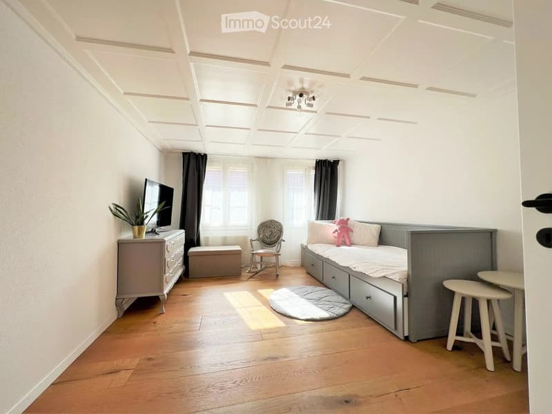 Cozy, well-lit room with wooden flooring, a bed with a headboard, a rocking chair, a small table with two stools, a television on a dresser, and a plant. The room features large windows with curtains and a decorative ceiling.