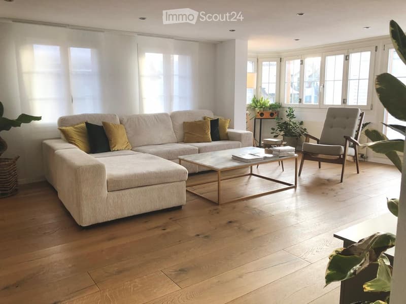 Bright living room with wooden flooring, large windows, a beige sectional sofa, a coffee table, and a wooden chair.