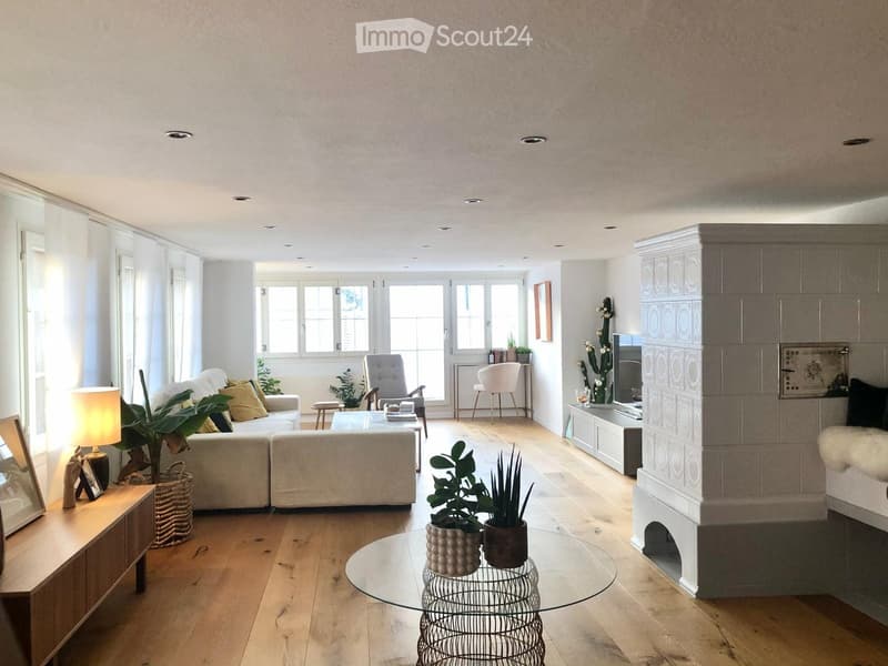 Bright living room with wooden flooring, large windows, a fireplace, and modern furniture including a sofa, coffee table, armchair, and potted plants.