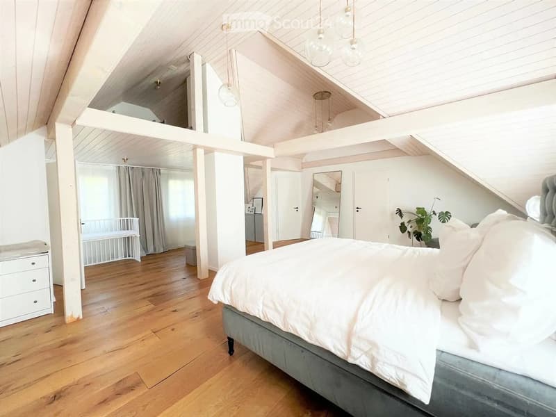 Bright, airy bedroom with wooden floors, white walls, and exposed wooden beams. Features a large bed with white bedding, a nightstand, a mirror, a small plant, and a baby crib in the background.