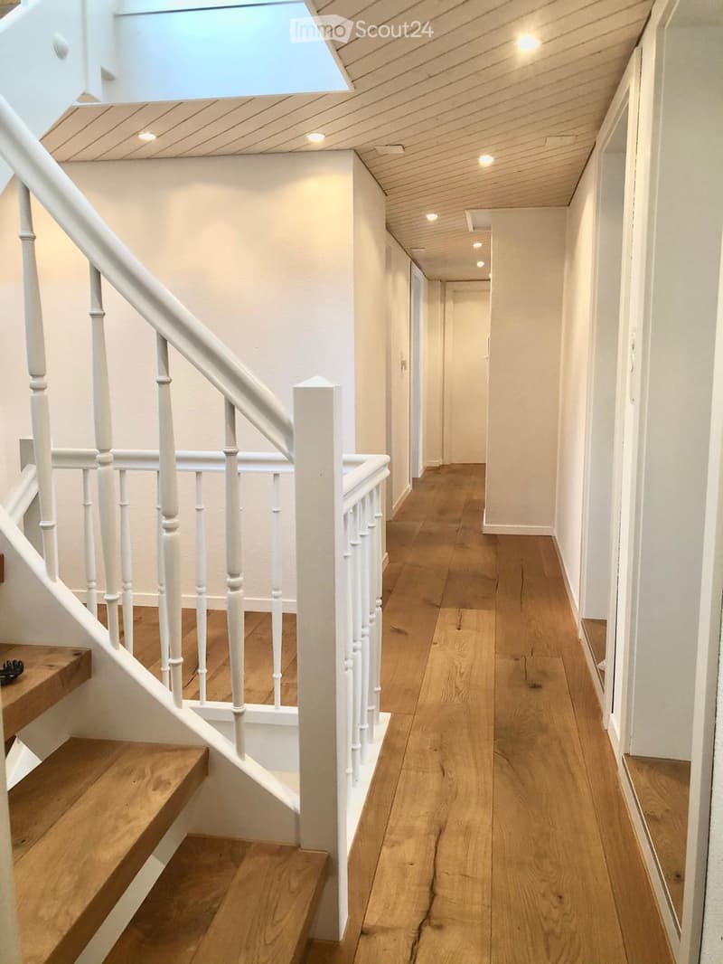 A well-lit hallway with wooden flooring, white walls, and a staircase with white railings leading upwards. The ceiling has recessed lighting and a skylight.
