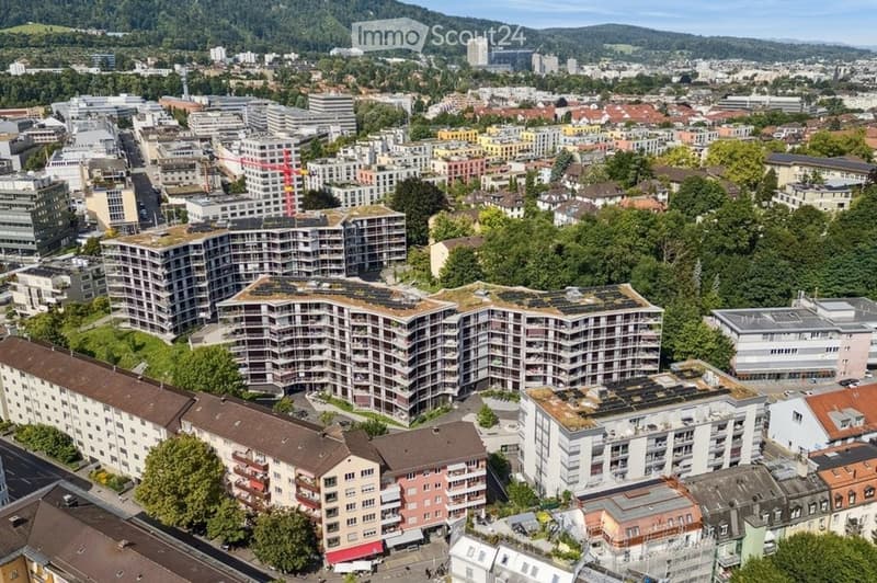 Aerial view of a cityscape with multiple residential and commercial buildings, green spaces, and a mountainous backdrop.