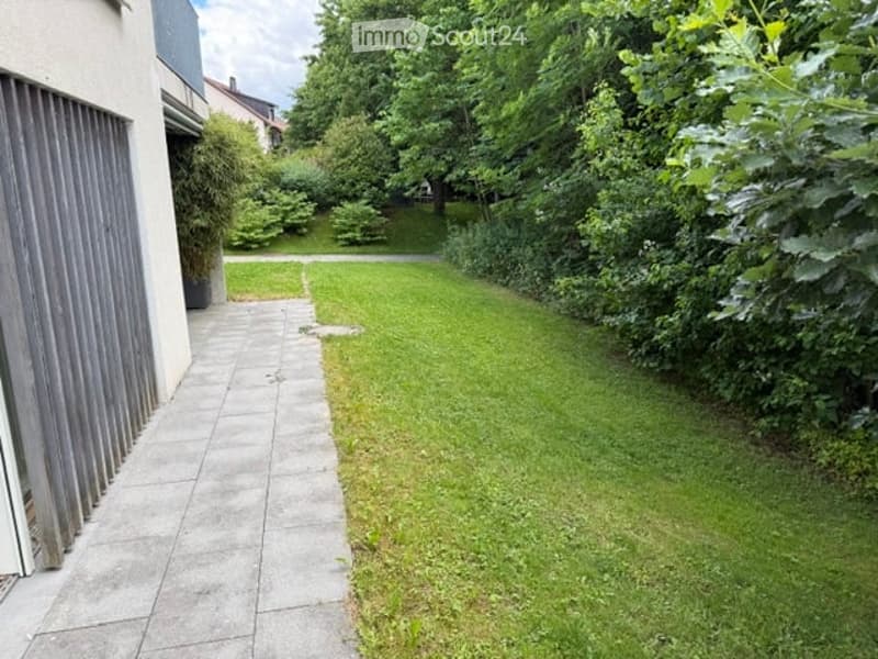 A well-maintained garden area with a paved pathway leading to a grassy lawn, surrounded by trees and shrubs. The garden is enclosed by a building on one side and dense greenery on the other.