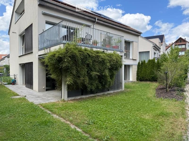 Modern two-story house with a balcony, green lawn, and surrounding trees.