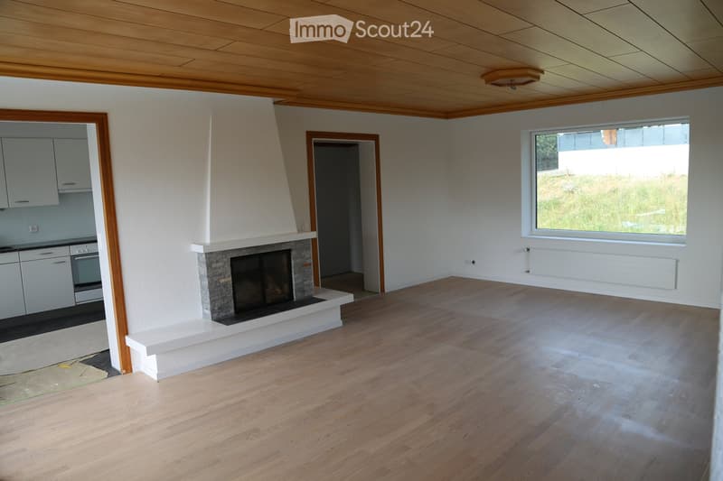 Empty room with wooden flooring, white walls, and a fireplace. The room has large windows and a visible kitchen area with cabinets and appliances.