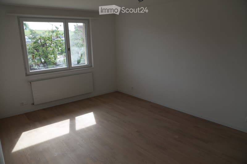 Empty room with wooden flooring, large window, radiator, and natural light streaming in.