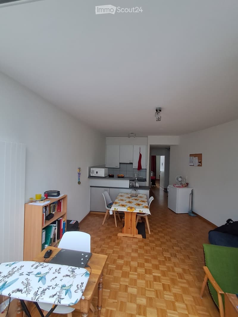 A cozy, well-lit room with wooden flooring, featuring a small kitchen area with cabinets, a microwave, and a refrigerator. There is a dining table with chairs, a bookshelf with books and a laptop, and a green armchair. The room also includes a fan and a s