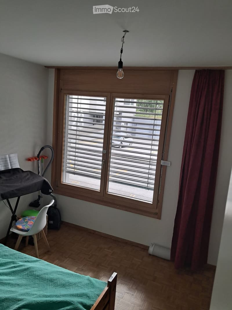 Bedroom with wooden flooring, large window with blinds, bed with green patterned bedspread, white chair, and vacuum cleaner in the corner.