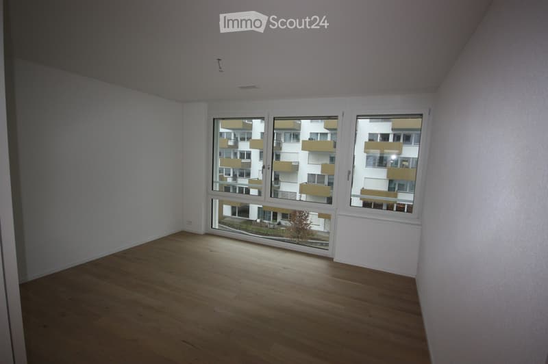 Empty room with wooden flooring and large windows showing a view of neighboring buildings and a small garden area.