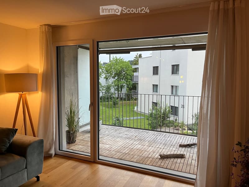 Living room with wooden flooring, large sliding glass doors opening to a wooden balcony, sofa, floor lamp, and potted plant near the entrance.