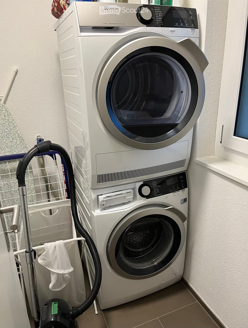 A compact bathroom with a stacked washing machine and dryer unit, a vacuum cleaner, and a laundry basket with a cloth.