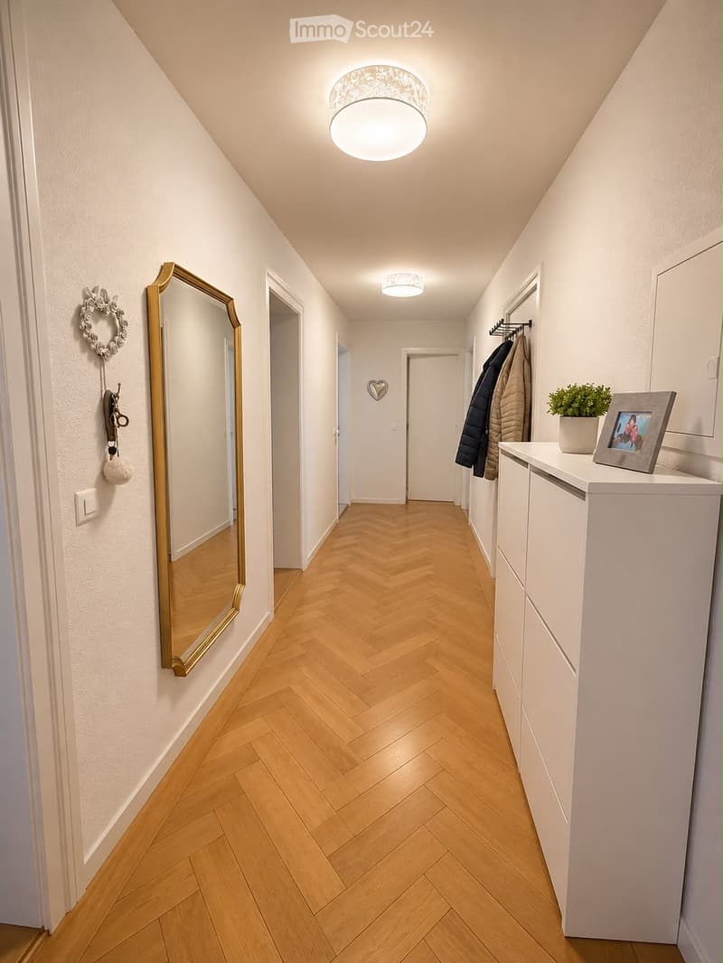 Hallway with herringbone wooden flooring, white walls, ceiling lights, coat rack, mirror, small table with plant and picture frame.