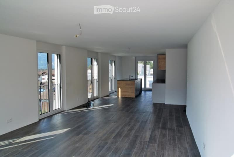 Empty room with dark wooden flooring, white walls, large windows, and a small kitchen area visible in the background.