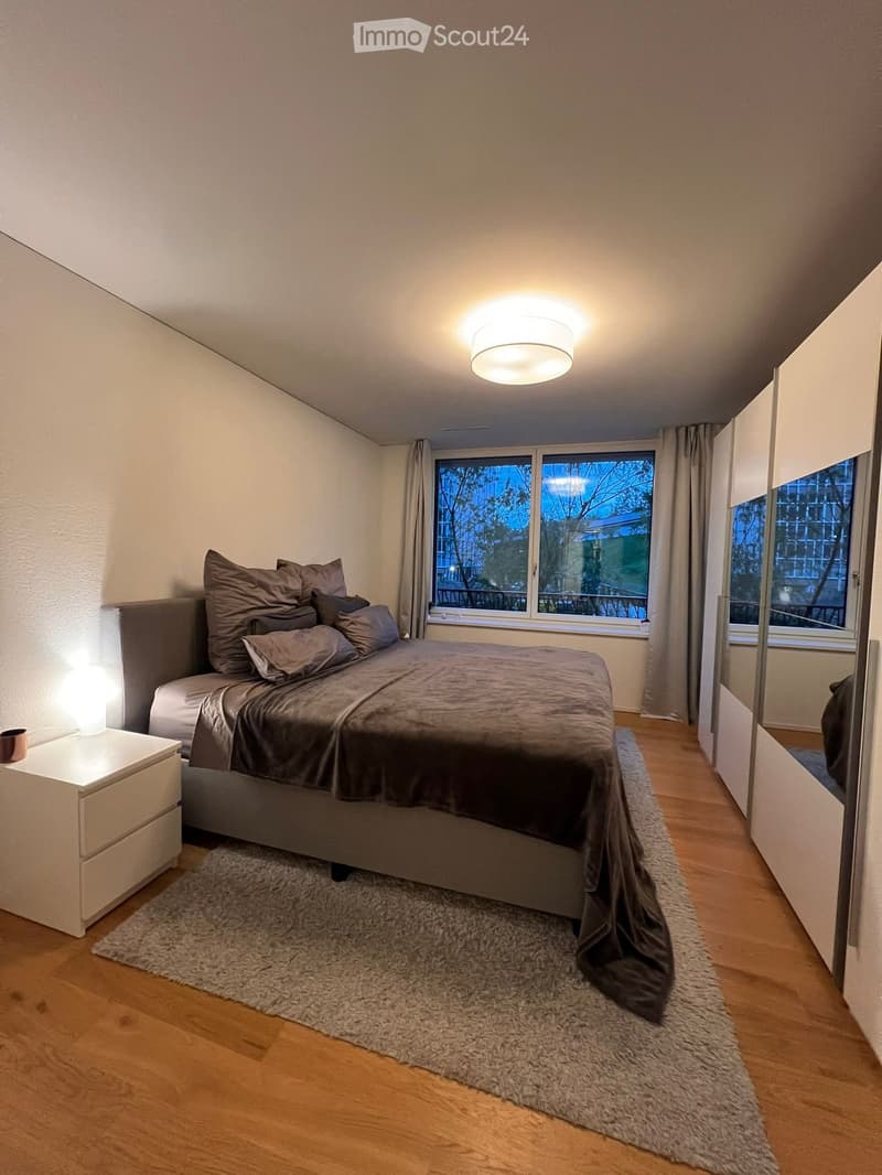 Modern bedroom with a large bed, nightstand, lamp, and large windows with curtains. The room features wooden flooring and a soft, grey rug.