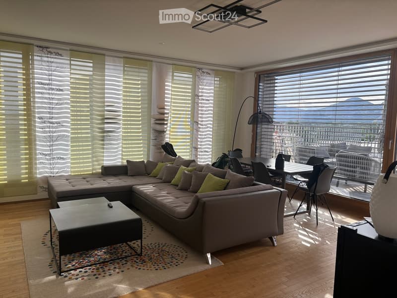 Modern living room with large windows offering a scenic mountain view, featuring a grey sectional sofa, coffee table, dining area with chairs, and a floor lamp.