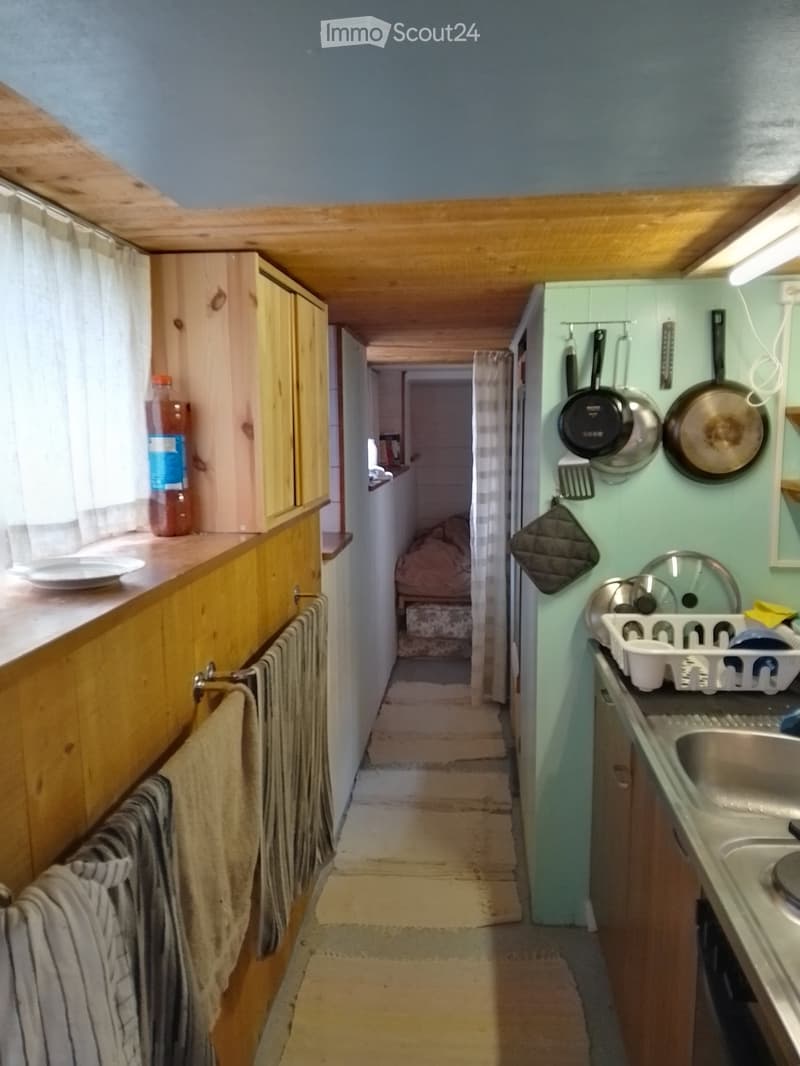 A narrow hallway with light blue walls, wooden ceiling, and a small kitchen area visible at the end. The kitchen features light green cabinets, a sink, and various kitchen utensils hanging on the wall.