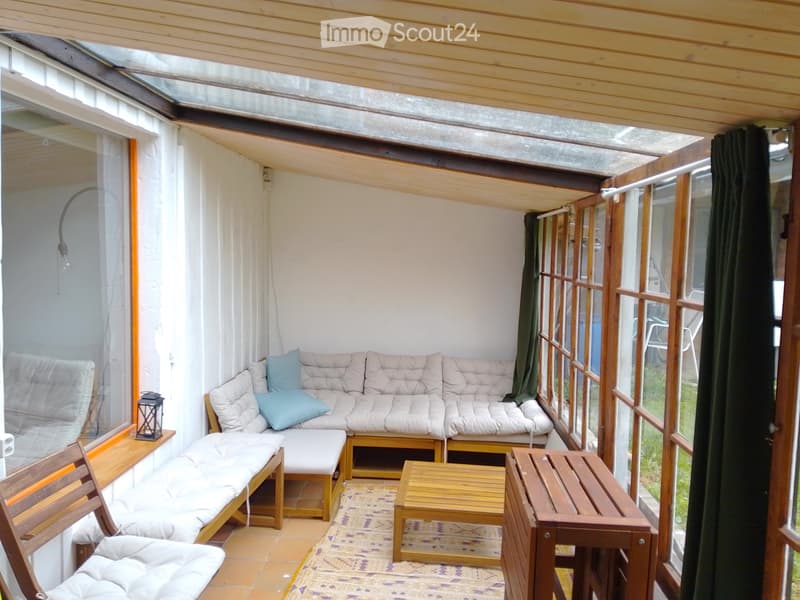 A cozy, enclosed balcony with a white sofa, wooden chairs, a small table, and a lantern-style lamp. The space features large windows and a wooden ceiling.